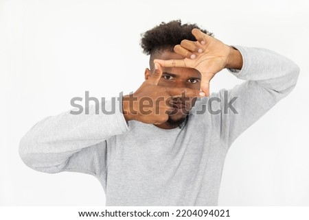 Portrait of concentrated African American man with frame gesture. Young bearded guy wearing white sweater peeping through finger frame against white background. Photographing concept
