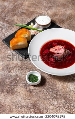 Ukrainian soup red borscht. From beets, cabbage, potatoes, pork, carrots and broth. Soup in a white, round, deep ceramic plate. The plate stands on a dark, coffee background. There is a slate, black, 