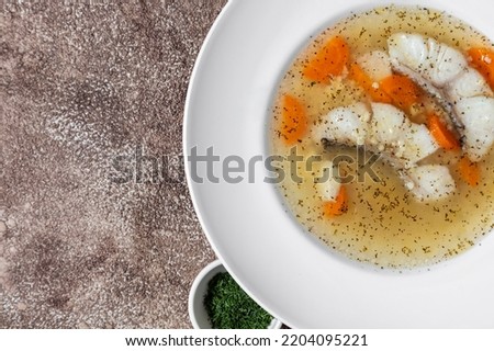 Fish soup Ukha from sturgeon. In a white, round, ceramic plate, there are two pieces of sturgeon fillet, chopped carrots and fish broth. Nearby is a small, white, ceramic bowl with finely chopped dill
