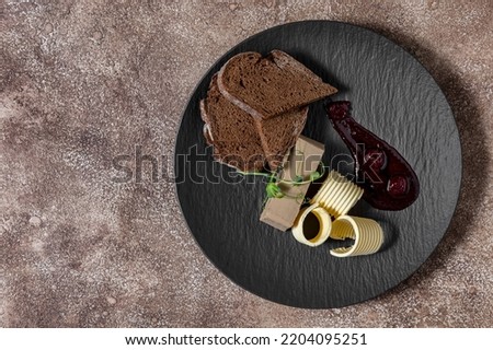 Rabbit pate with dark buckwheat toast, butter and cherry sauce. Microgreen sprouts lie on top of the pate. The food lies on a black, round, slate plate. The plate stands on a fabric background, coffee