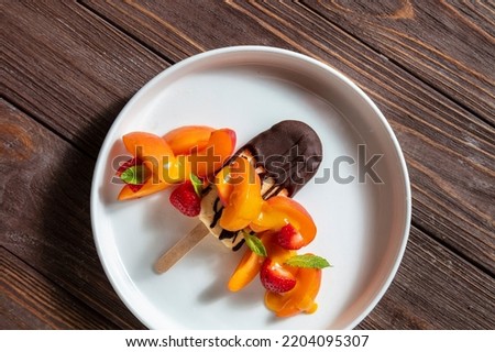 Ice cream on a stick in dark chocolate with peaches, strawberries, mint in a plate on a wooden table