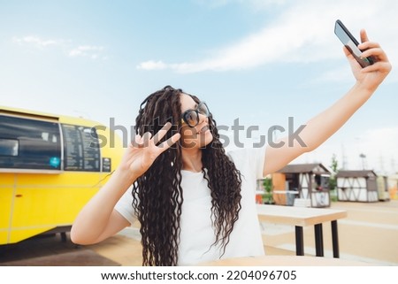 A laughing girl with dreadlocks is sitting in a street cafe and taking a selfie with her phone. a woman smiles and takes pictures of herself on a nice autumn day . generation z