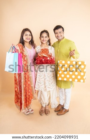 Happy young indian family holding gift box and shopping bags celebrating diwali festival together isolated on studio background. festive shopping and sale, looking at camera.