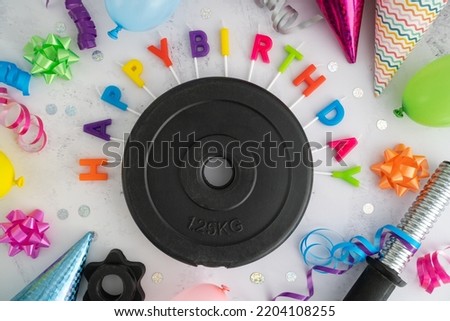 Dumbbell barbell weight plates with colorful Happy Birthday cake candles letters, balloons, cone hats, bows and ribbons. Gym exercise equipment as a gift idea for birthday party, flat lay composition.