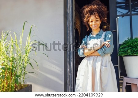 Cafe owner standing in the doorway of her coffee shop. Cheerful young pretty female barista standing at the door of a restaurant and looking at camera.