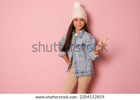 Pretty brunette kid girl wearing white winter hat over pink background Doing peace symbol with fingers, smiling cheerful showing victory