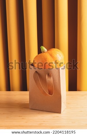 Orange pumpkin in kraft shopping bag on wooden table against yellow fabric curtains, studio shot. Halloween holiday, autumn shopping, fall season concept.