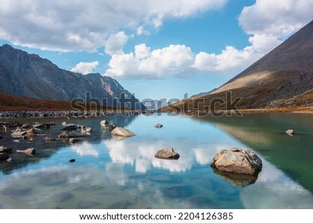 Tranquil autumn landscape with clouds reflection on smooth mirror surface of mountain lake in high hanging valley. Meditative view from calm alpine lake to mountain vastness. Stones in clear water. Royalty-Free Stock Photo #2204126385