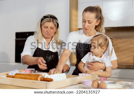 Adorable young mother wearing apron holding little daughter while kneading dough for christmas cookies teaching young girl cooking baking. Granny helping managing process.