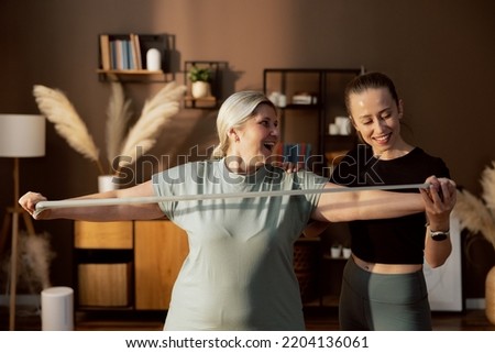 Middle aged woman looking at young caregiver stretching together using resistance band wearing sportswear doing exercises sports standing indoors.