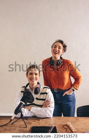 Two female podcasters smiling at the camera with headsets around their necks. Happy women co-hosting an audio broadcast in a home studio. Cheerful content creators recording an internet podcast.