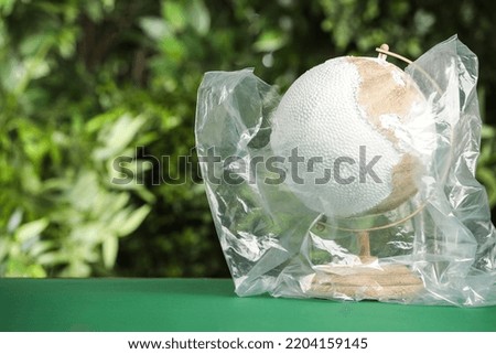 Globe in plastic bag on table against green leaves, space for text. Environmental conservation