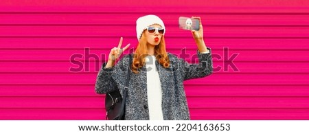 Portrait of stylish young woman taking selfie with smartphone wearing knitted white hat on colorful pink background