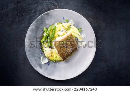 Traditional fried skrei cod fish filet with green asparagus tips and mashed potato creme in parmesan olive oil sauce as top view on a modern design plate with copy space 