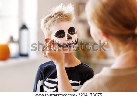 Festive makeup for Halloween. Woman doing skeleton make-up    for a little cheerful boy in costume while preparing holiday at home.
