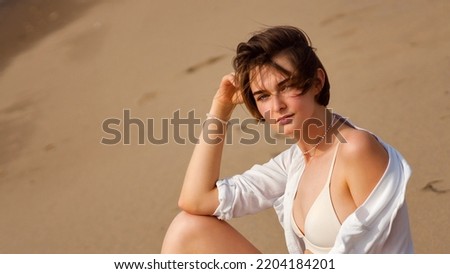 Close up portrait girl with short haircut wears white summer shirt and bikini swimsuit. Pensive young caucasian short hairdo woman sitting on sandy beach windy summer day. 