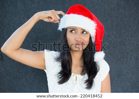 Closeup portrait of a cute Christmas woman with a red Santa Claus hat, white dress, finger on head, confused, thinking what to shop for the holiday season. Emotion on isolated grey background.
