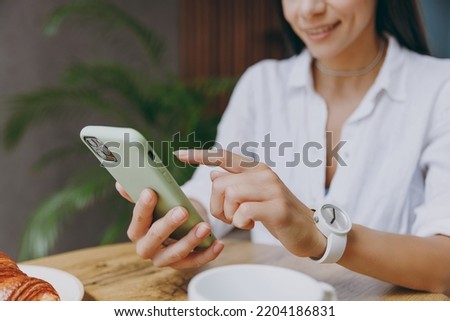Cropped close up photo smiling young latin woman 30s wear white shirt use mobile cell phone in green case sit alone at table in coffee shop cafe restaurant indoors. Freelance office business concept
