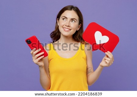 Young minded happy woman 20s she wear yellow tank shirt use mobile cell phone heart form like icon sign from social network media feedback look aside isolated on plain pastel light purple background