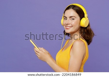 Side view young woman 20s wear yellow tank shirt headphones listen to music hold in hand use mobile cell phone look camera isolated on plain pastel light purple background. People lifestyle concept Royalty-Free Stock Photo #2204186849