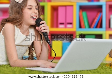 A teenage girl with a laptop sings into a microphone lies on the carpet