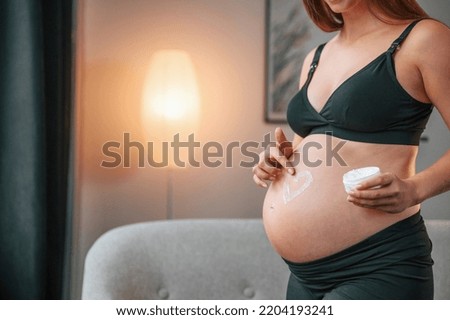 Making heart shaped picture by cream on the belly. Beautiful pregnant woman is indoors at home.