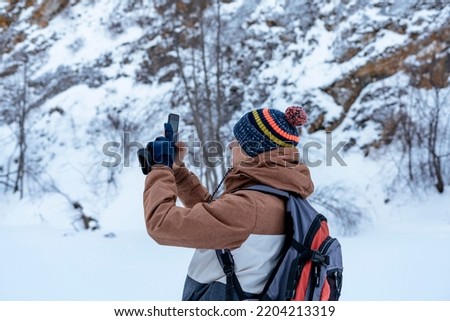 young man in brown warm clothes with backpack walking in snow among rocks and cliffs in winter looking at view taking photos with smartphone Active lifestyle hiking selective focus