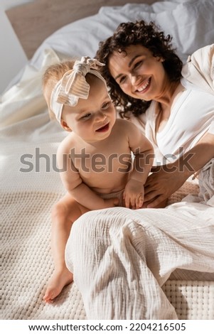 happy woman with curly hair lying on bed and looking at infant daughter in headband