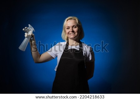 A blond gardener woman in a black apron and protective glove holds a sprayer in her hands. Dark background.
