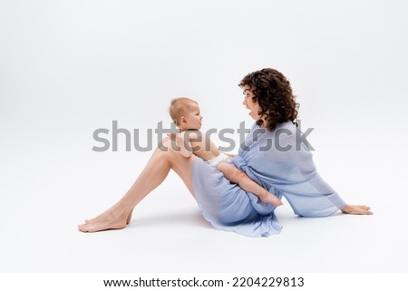 Side view of shocked mother in dress looking at baby girl in panties on white background