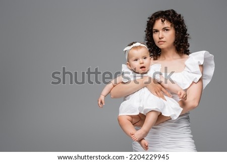 Curly woman in white dress holding baby girl isolated on grey