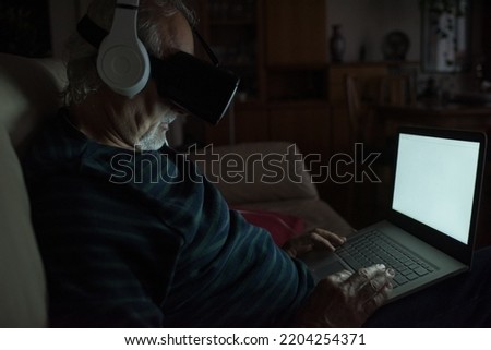 Senior adult man indoors at home sitting couch using laptop and 3d goggles