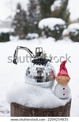 model of a toy metal excavator stands on a mirror disco ball, a souvenir snowman on a snow-covered stump. concept of christmas business greetings, winter holidays in construction companies