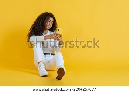 Relaxed young woman sitting on floor using her cell phone on yellow background