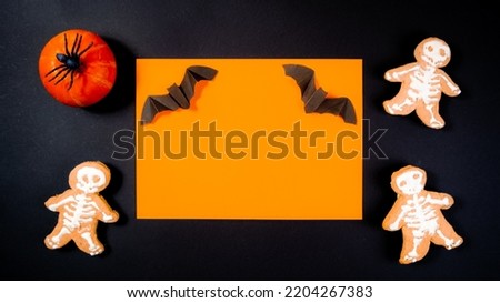 Bat on orange background in black rectangular frame adorned with a small orange pumpkin with a black spider and skeleton-shaped gingerbread. Halloween holiday concept.