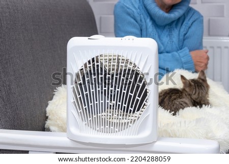 A heating fan heats a woman sitting on a bed with a cat, the woman is covered with a plaid. Concept: a cold snap in the apartment, heating shutdown, rising gas prices. Royalty-Free Stock Photo #2204288059