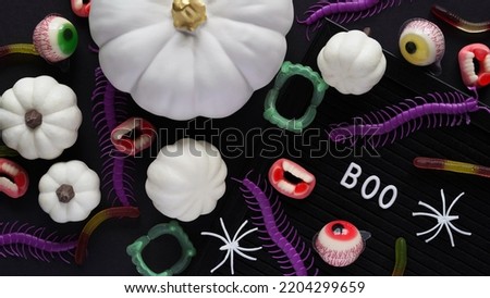 Pumpkins, cauldrons, pots, worms, jaws, spiders, eye-shaped candies and a text board with the inscription BOO on a black background, top view. A collection of items for a Halloween party