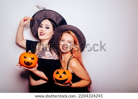 Two girls in witch costumes on a white background hold pumpkins, smile and laugh for Halloween. Autumn photo of girlfriends from a costume party