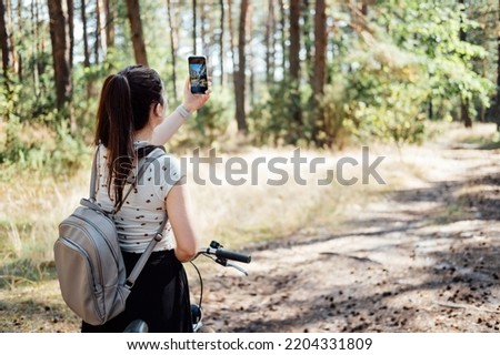 Young woman with backpack riding bike and taking photo on cell phone on pine forest background. Girl with bike shooting photo by phone camera in forest Royalty-Free Stock Photo #2204331809