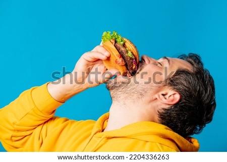 Happy white guy in yellow hoodie eating hamburger on blue background