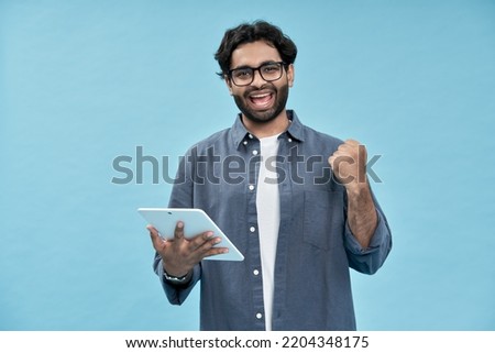 Happy young arab man student winner winning online raising fist in yes gesture using digital tablet screaming winning online celebrating getting new job isolated on blue background.