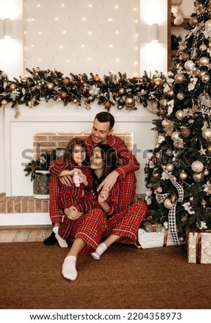 Dad, mom and their daughter in red pajamas sit near the Christmas tree and fireplace on Christmas Eve. Happy young family having fun enjoying the Christmas holidays. Beautiful Christmas decor