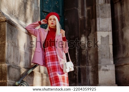 Fashionable elegant blonde woman wearing stylish red beret, turtleneck, classic pink suit blazer, checkered skirt, holding trendy bag, posing in street of European city. Copy, empty space for text Royalty-Free Stock Photo #2204359925