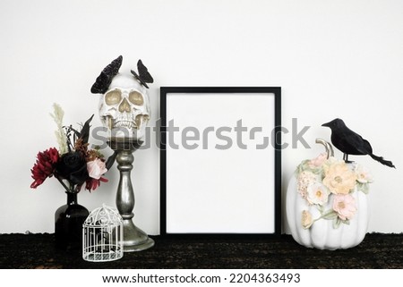 Halloween gothic romance mock up. Black frame on a black shelf with red and black flowers, skull and floral pumpkin. Portrait frame against a white wall. Copy space.