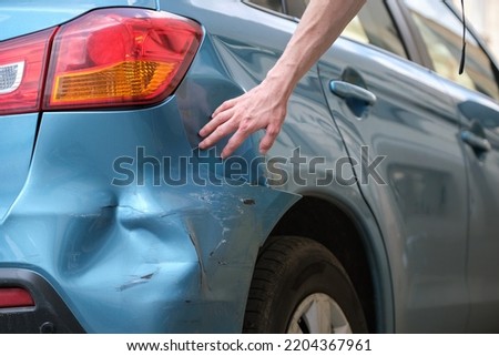Driver hand examining dented car with damaged fender parked on city street side. Road safety and vehicle insurance concept Royalty-Free Stock Photo #2204367961