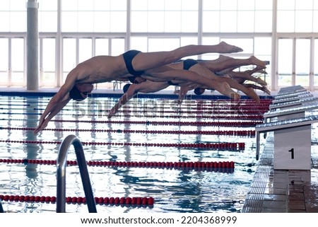 Side view of multi-ethnic group of male swimmers at swimming pool, jumping from starting blocks, plunging into water. Swimmers training hard for competition. Royalty-Free Stock Photo #2204368999