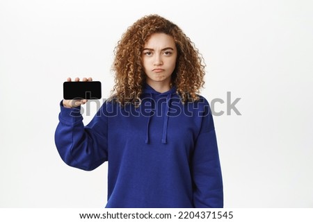 Angry, upset curly girl, showing horizontal smartphone screen, mobile phone app, frowning and sulking grumpy, complaining, feeling jealous, standing over white background