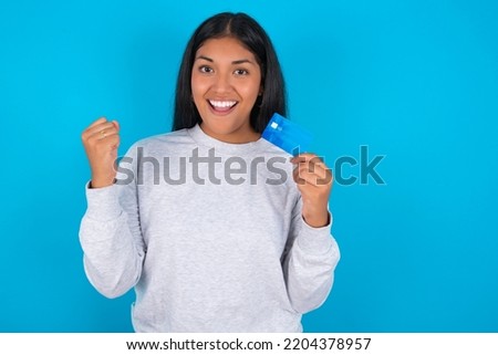 Photo of lucky impressed young latin woman wearing grey sweater over blue background arm fist holding credit card. Celebrated