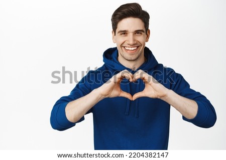 Handsome young man smiles and shows hand heart sign, I love you gesture, making romantic confession, standing over white background.