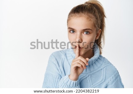 Close up portrait of beautiful little teen girl, shows hush, taboo quiet gesture, tell secret, press one finger to lips and say shh, stands in winter sweater over white background.